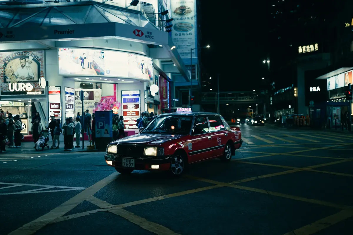 A classic red taxi navigates the bustling streets of Hong Kong at night under neon lights.